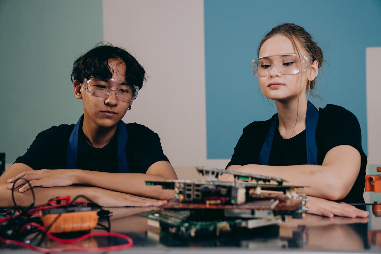 menu-12 Two young engineers wearing safety goggles focus on an electronic project indoors.